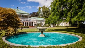 Una casa grande con piscina y fuente, rodeada de césped y árboles bien cuidados.
