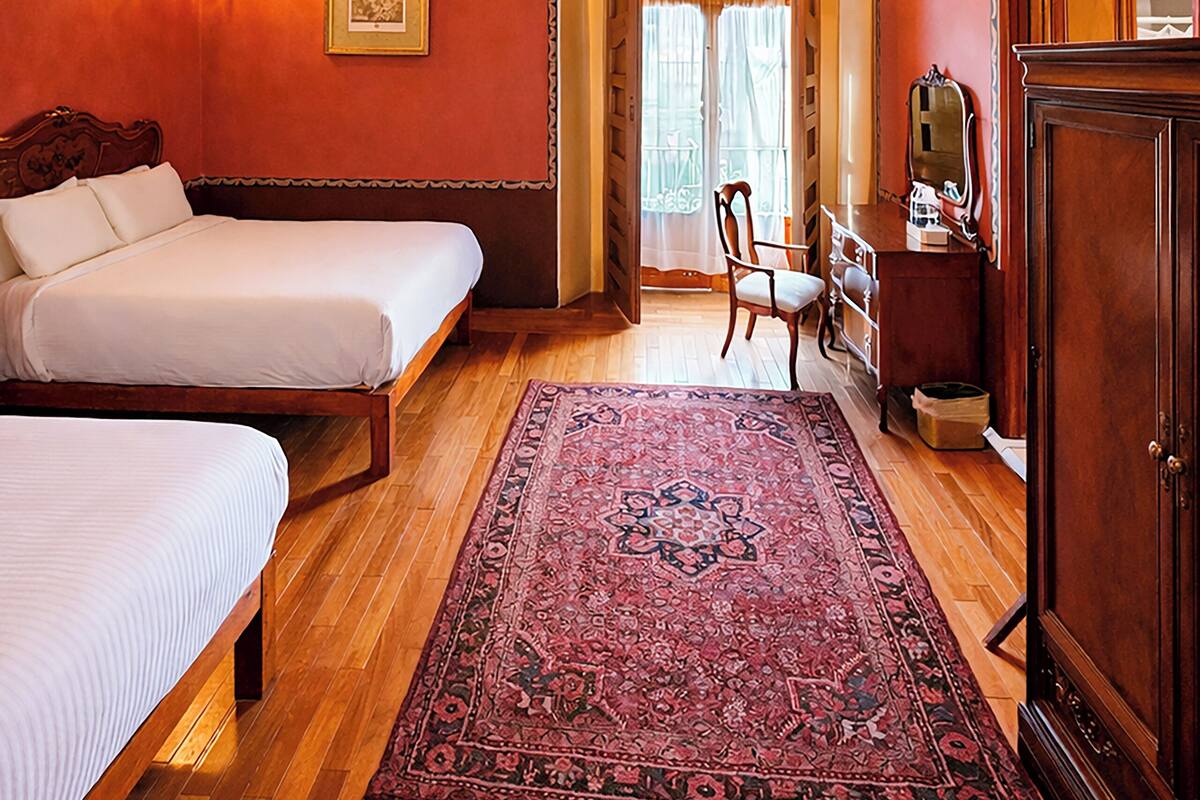Un dormitorio con techo de madera, candelabro, cama, sillón, cómoda y espejo.