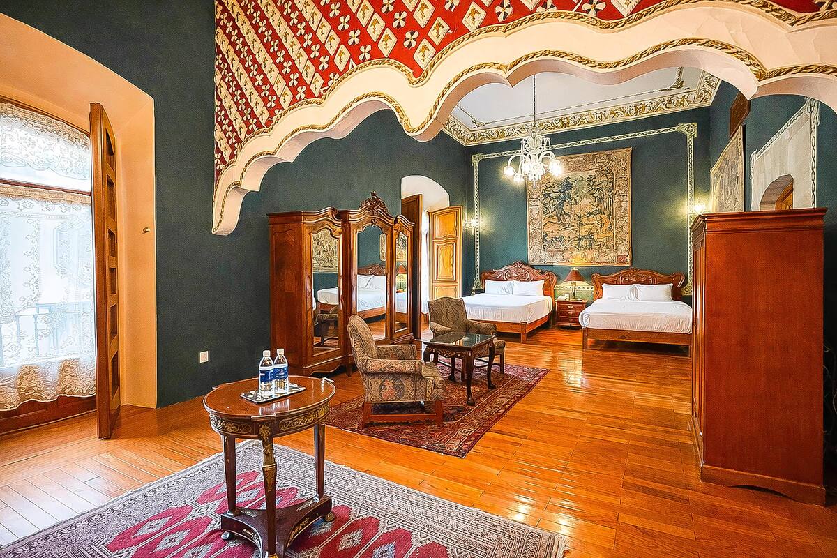 Habitación de hotel con una cama grande, dos camas más pequeñas, una zona de estar con sillas, una mesa y una araña.