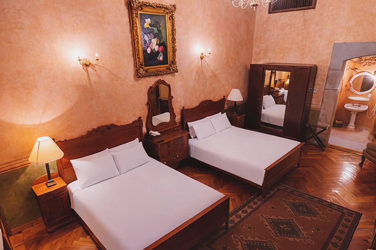 Habitación de hotel con dos camas, suelo de madera, candelabro, un cuadro y baño con espejo y lavamanos.