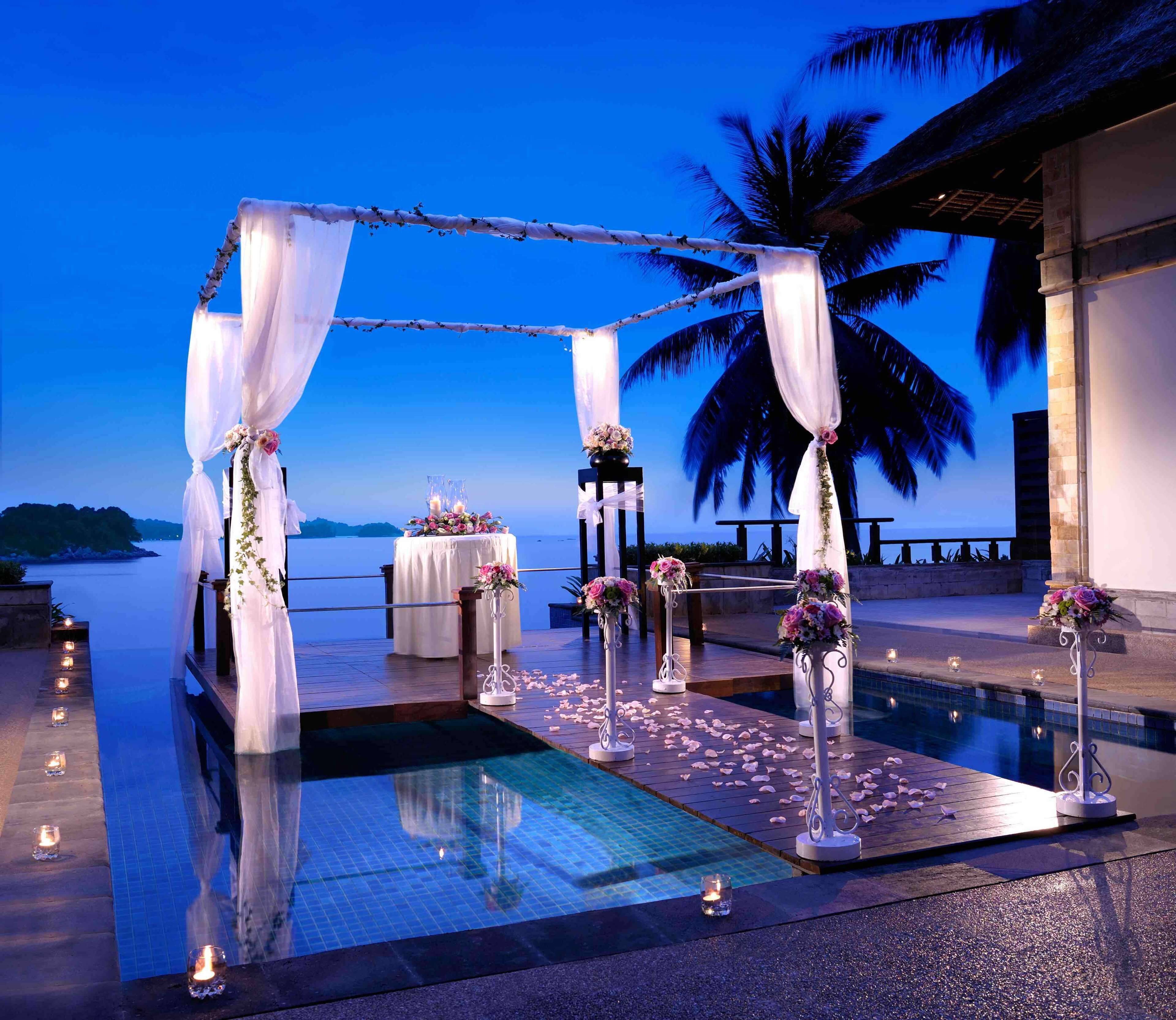 A wedding ceremony setup with a pool, palm trees, and a view of the sea.
