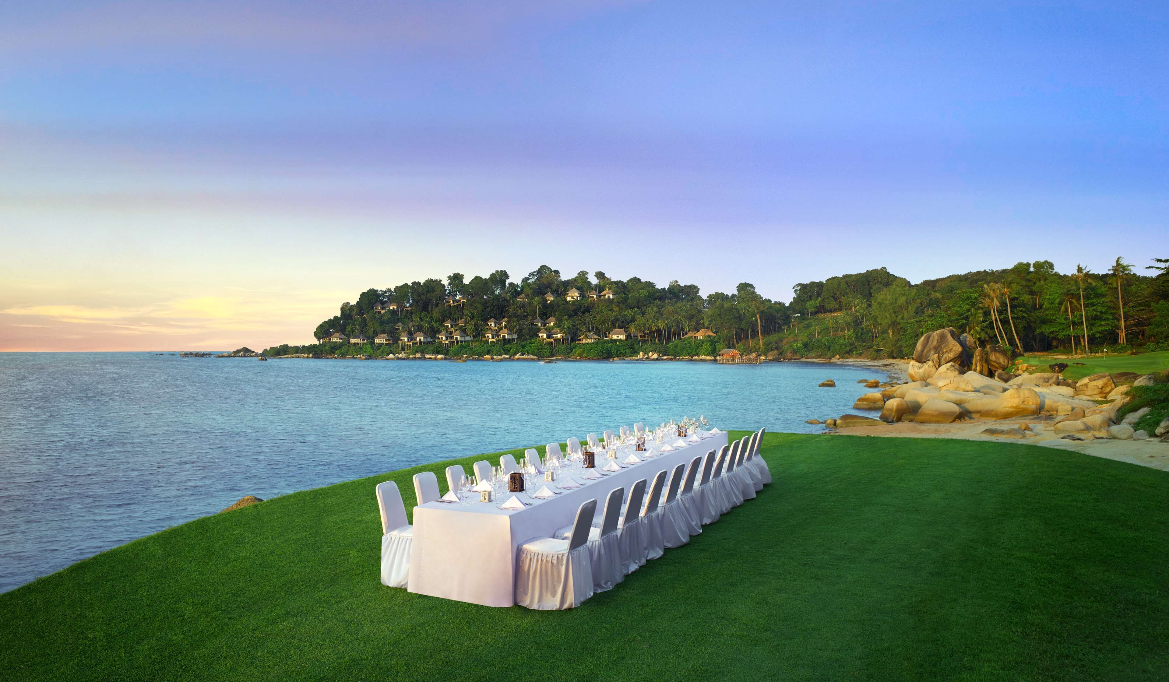 Una larga mesa dispuesta para un evento formal, con vistas a un mar tranquilo y una colina boscosa al fondo.