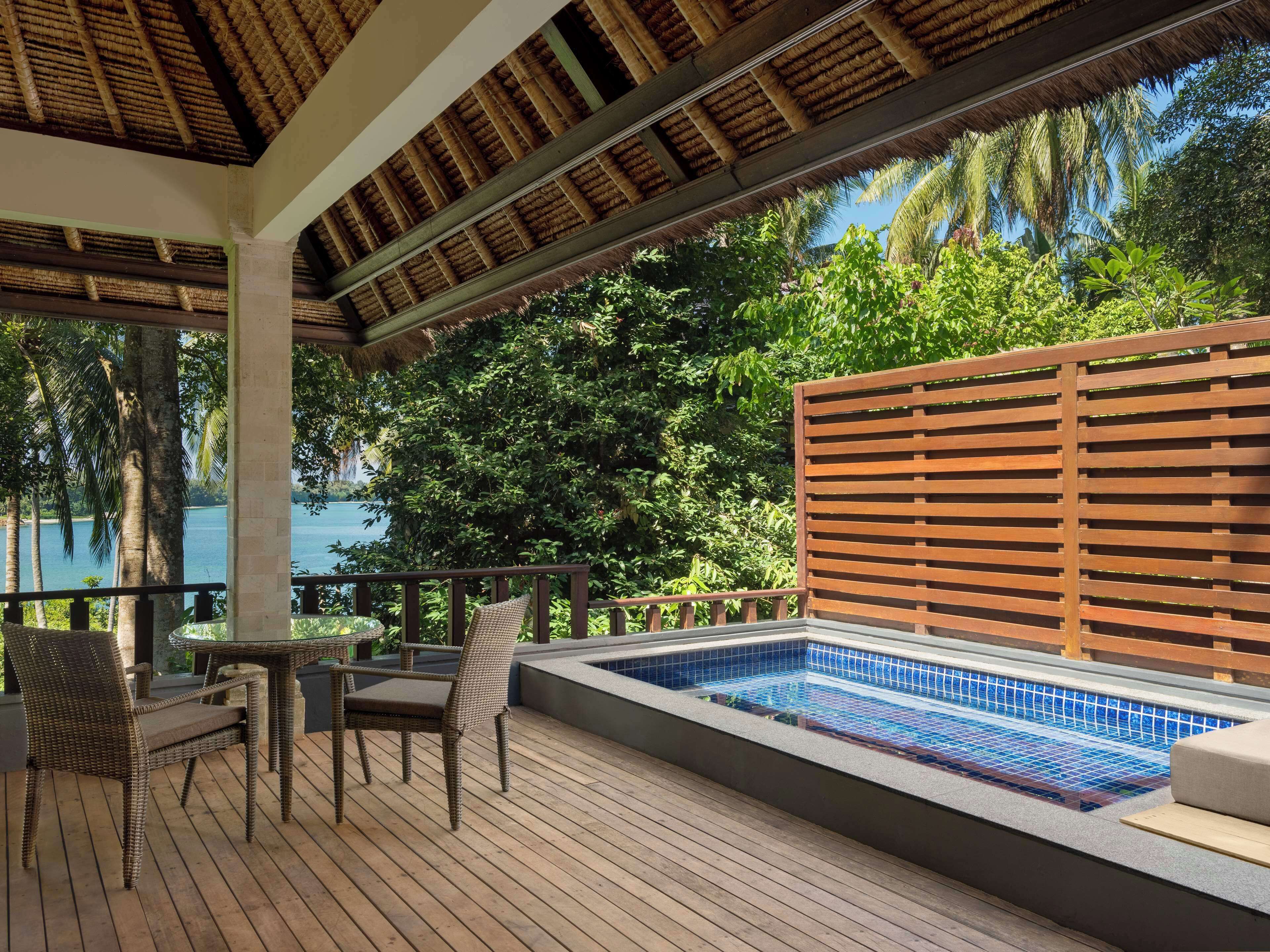 Ein überdachter Außenbereich mit Pool, Holzterrasse, Korbmöbeln und Blick auf ein Gewässer.