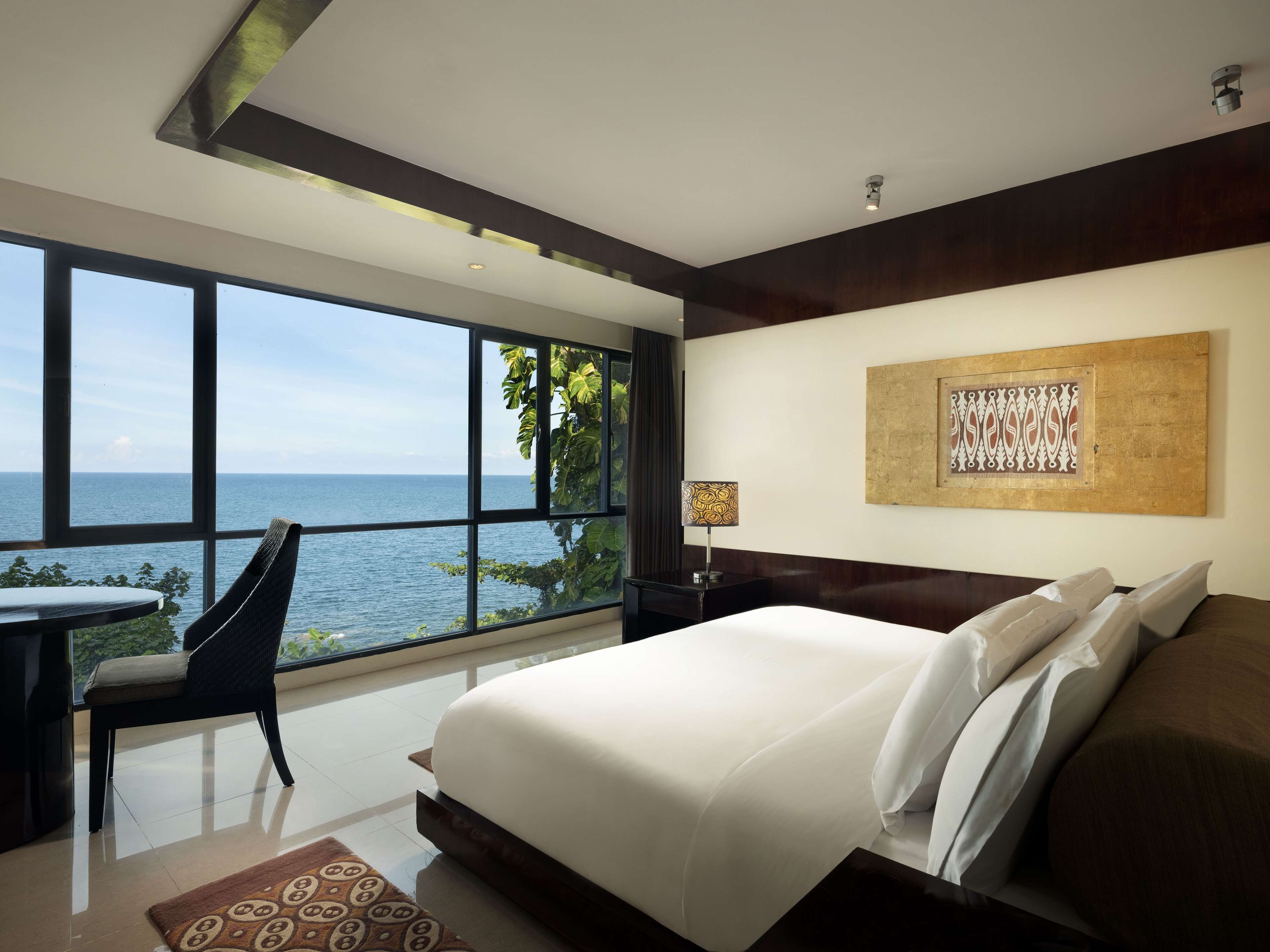 A hotel room with a large window offering a sea view, a bed with white linens, a desk with a chair, and a wall-mounted lamp.