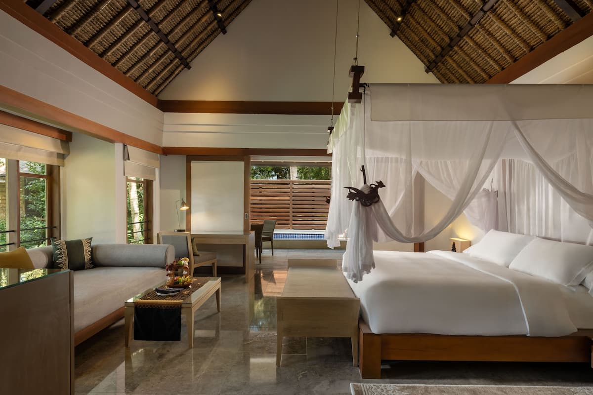 A spacious bedroom with a large bed, a seating area with a sofa and a table, and a view of the outdoors through large windows.