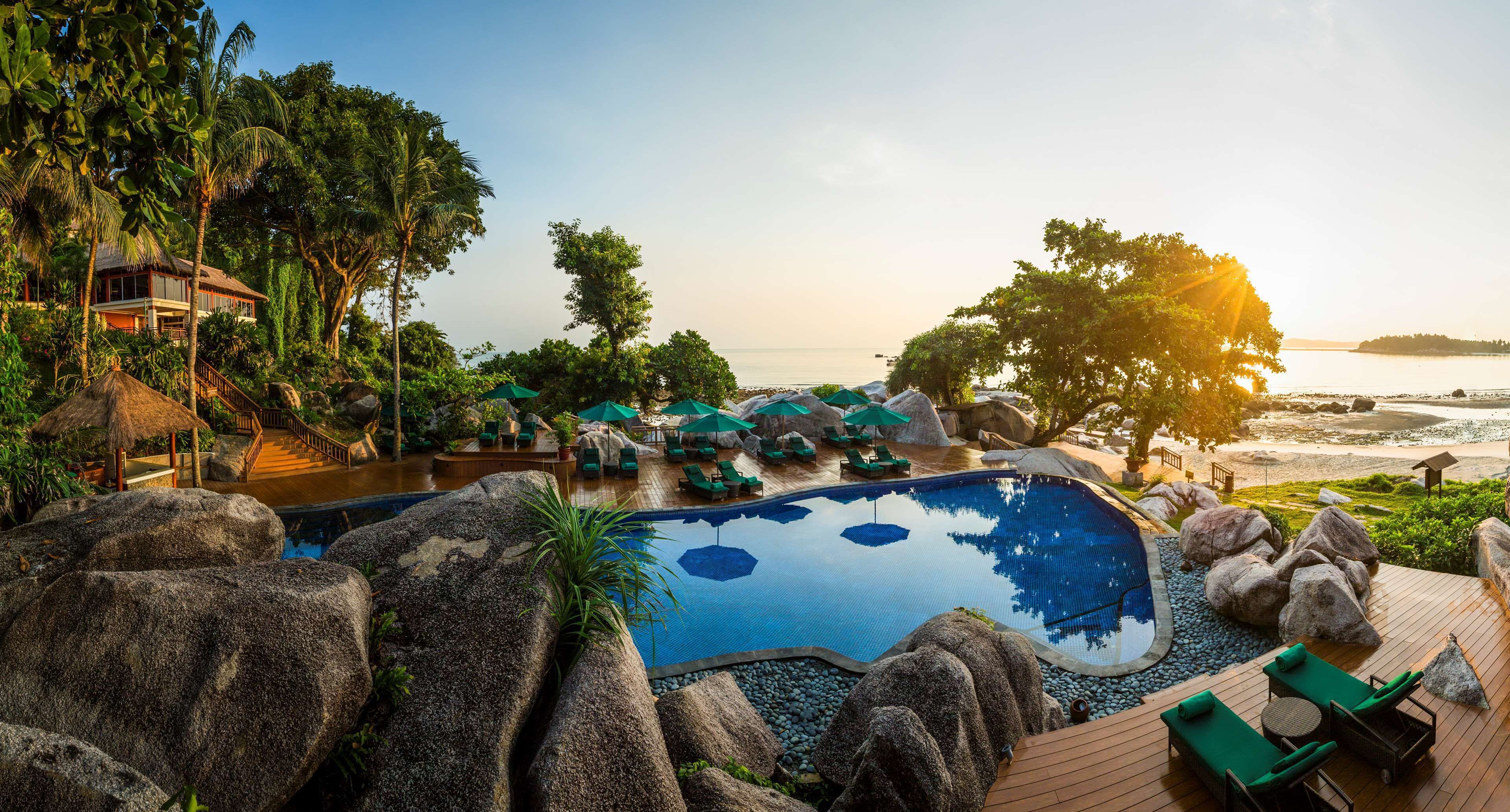 Ein Poolbereich in einem Resort mit Blick auf Strand und Meer, umgeben von Felsen und Grünpflanzen.