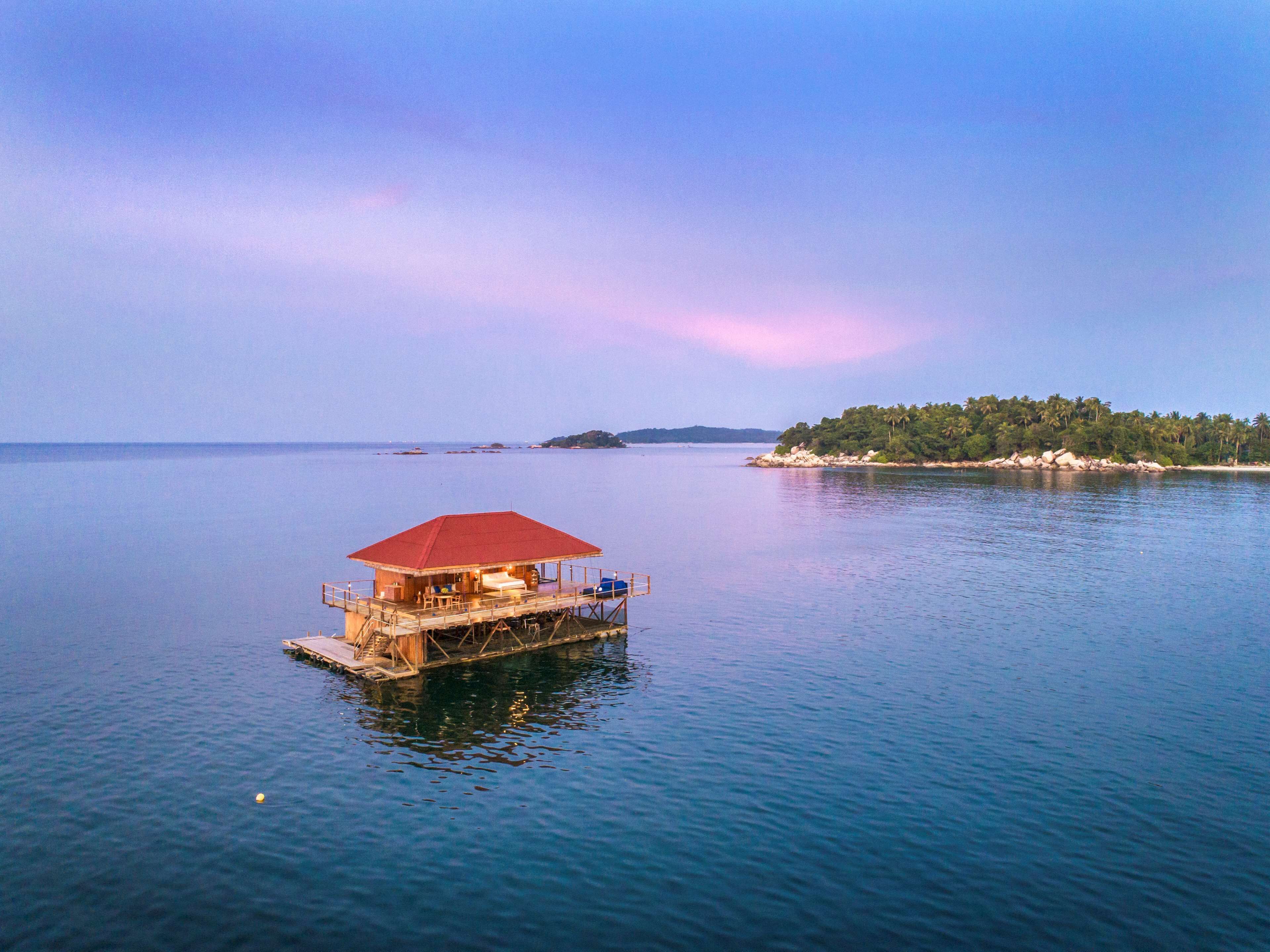 A floating structure with a red roof, a deck, and a small pool, surrounded by calm water and a distant island with vegetation.