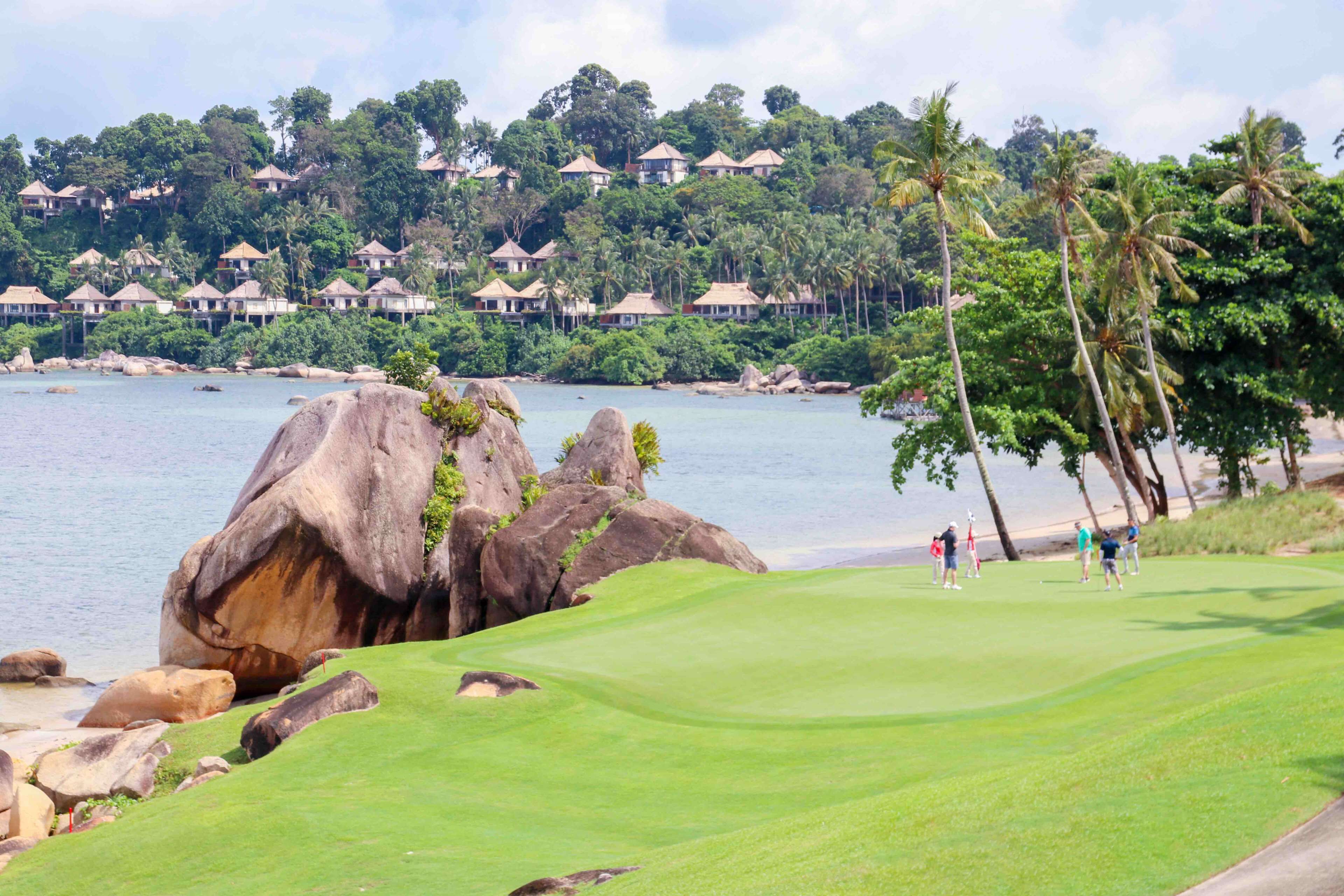 Un campo de golf con una gran formación rocosa, un cuerpo de agua y edificios de un resort al fondo.