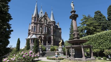 A historic building with a prominent tower and statues, surrounded by well-maintained gardens and a fountain.
