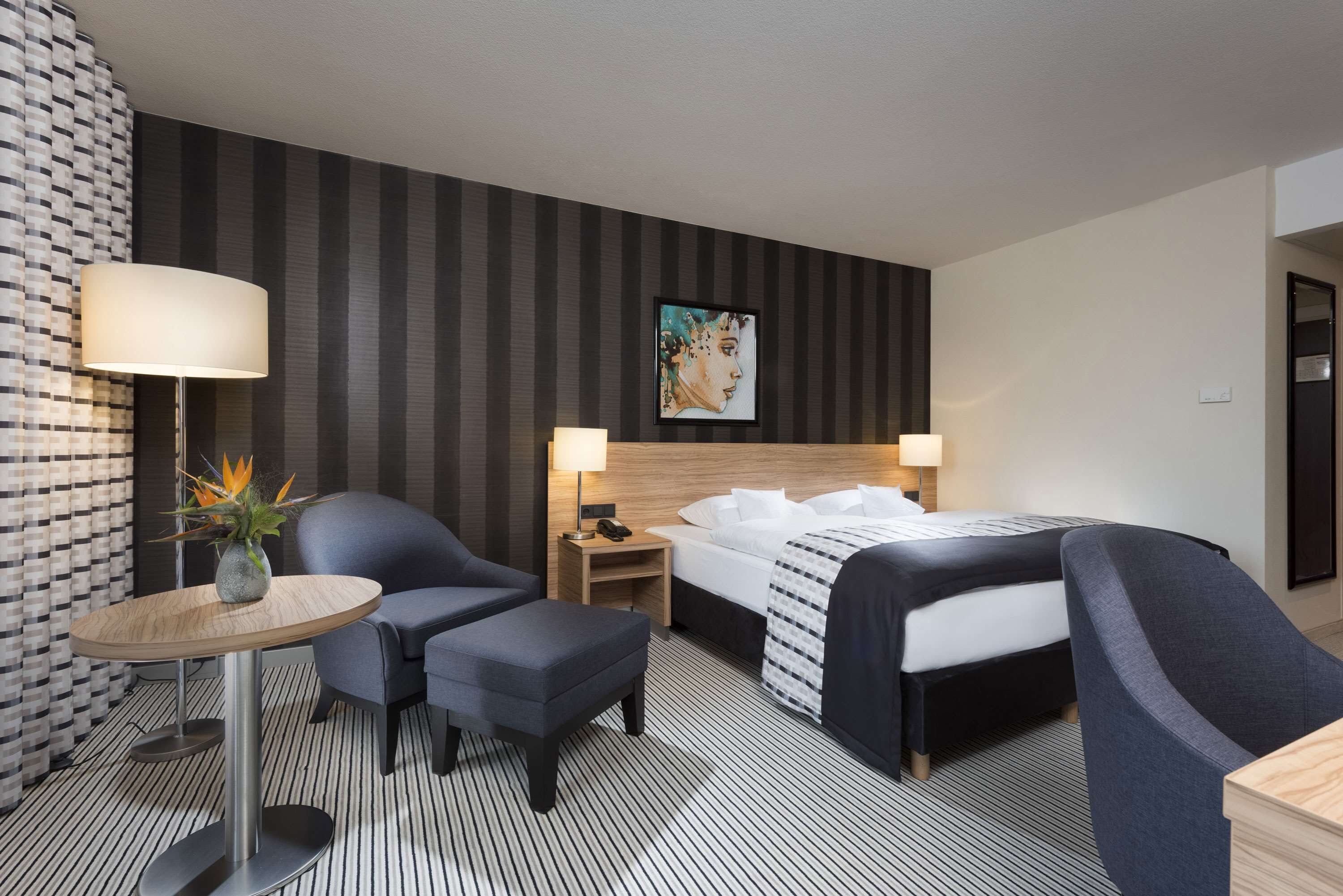 Habitación de hotel con cama, dos sillones, una mesita con lámpara, una mesita de noche con lámpara y una pared con estampado a rayas y un cuadro enmarcado.