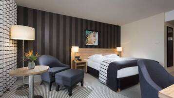 Habitación de hotel con cama, dos sillones, una mesita con lámpara, una mesita de noche con lámpara y una pared con estampado a rayas y un cuadro enmarcado.