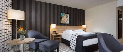 Une chambre d’hôtel comprenant un lit, deux chaises, une petite table avec une lampe, une table de chevet avec une lampe, et un mur orné d’un motif à rayures et d’un tableau encadré.