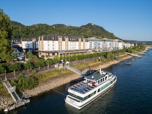 Un hotel frente al río con una barca amarrada, un puente y una colina con edificios.