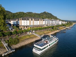 Un hotel frente al río con una barca amarrada, un puente y una colina con edificios.