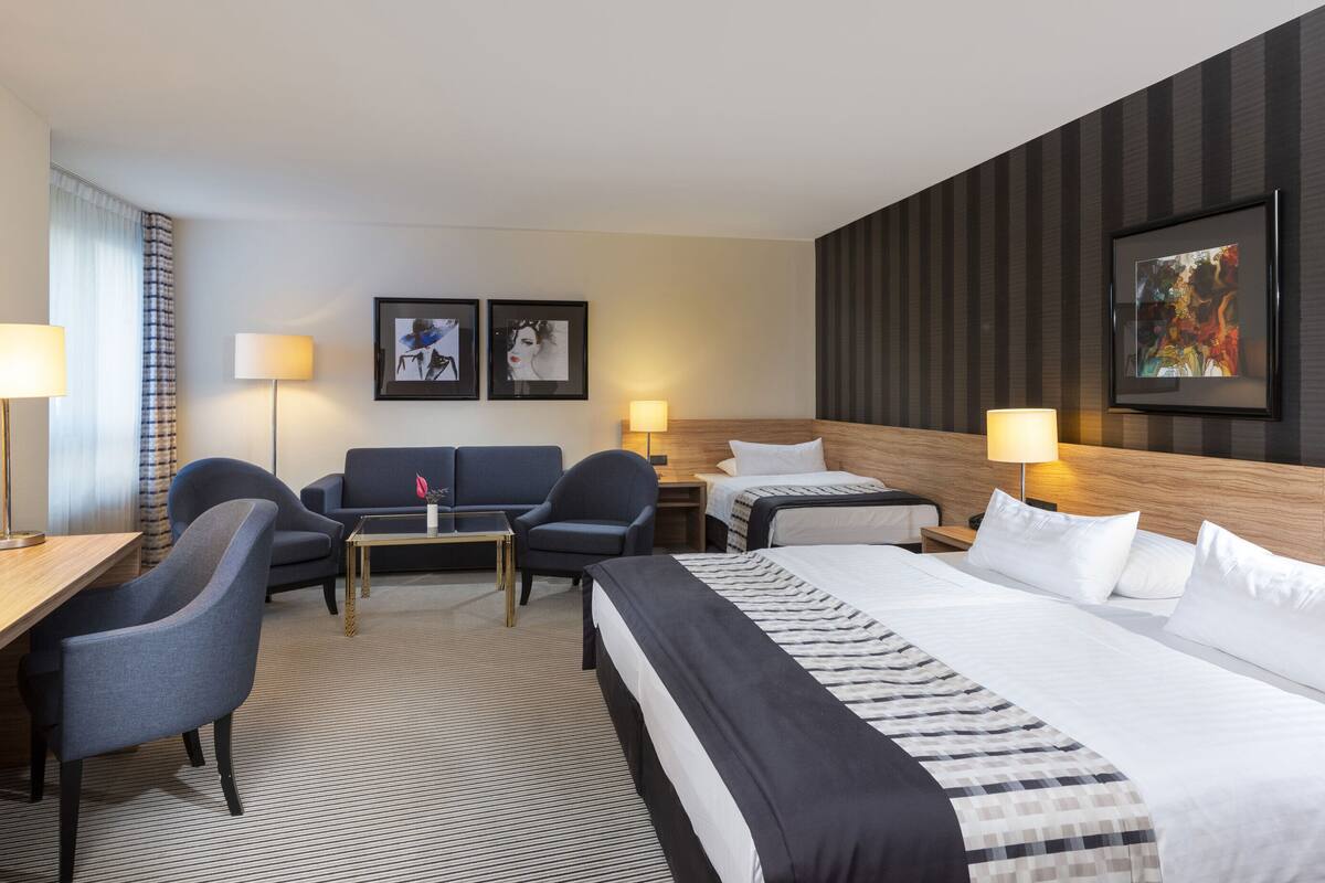 A hotel room with a large bed, two armchairs, a sofa, a desk, and a flat-screen TV. There are two framed artworks on the wall and a lamp on each side of the bed.