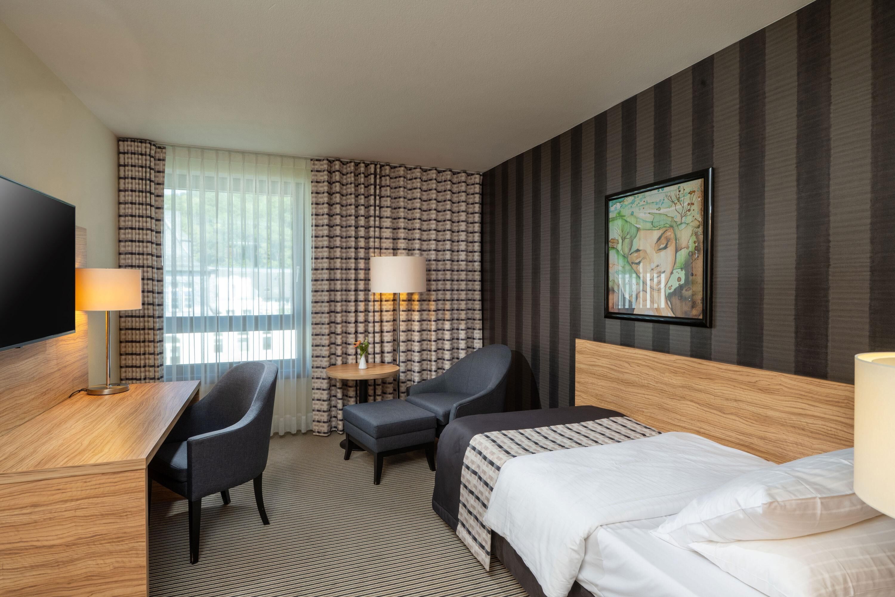 A hotel room with a bed, two chairs, a desk, a TV, and a painting on the wall.