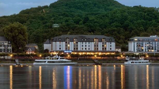 A waterfront hotel with a large building, boats docked in front, and a hill in the background. - Maritim Hotel Königswinter (Königswinter)