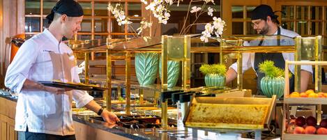 Montaje tipo buffet con varias estaciones de comida, un mostrador con vitrina de vidrio y detalles decorativos como flores.