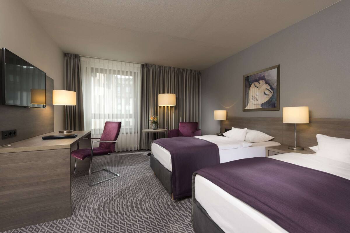Ein Hotelzimmer mit zwei Betten, einem Schreibtisch, einem Sessel, einem Fernseher und einem Bild an der Wand.