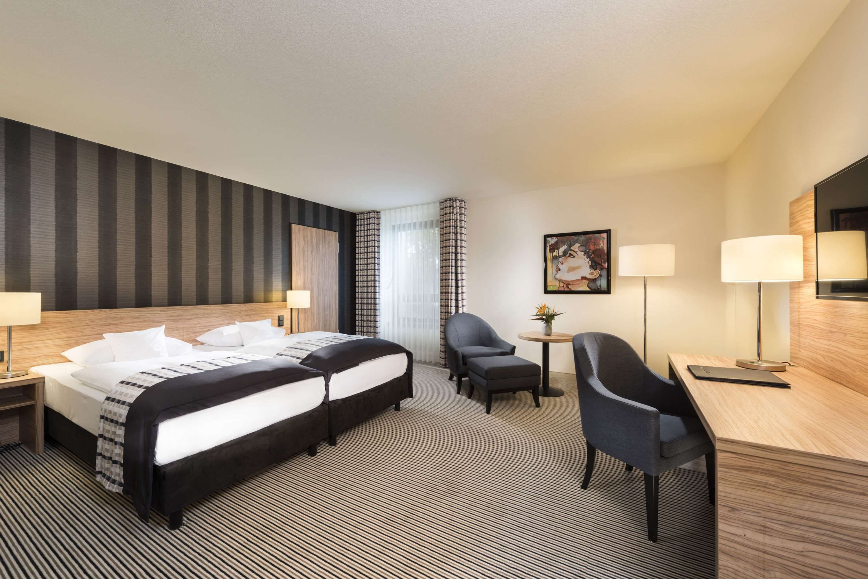 A hotel room with two beds, a desk, a chair, a small table, and a painting on the wall.