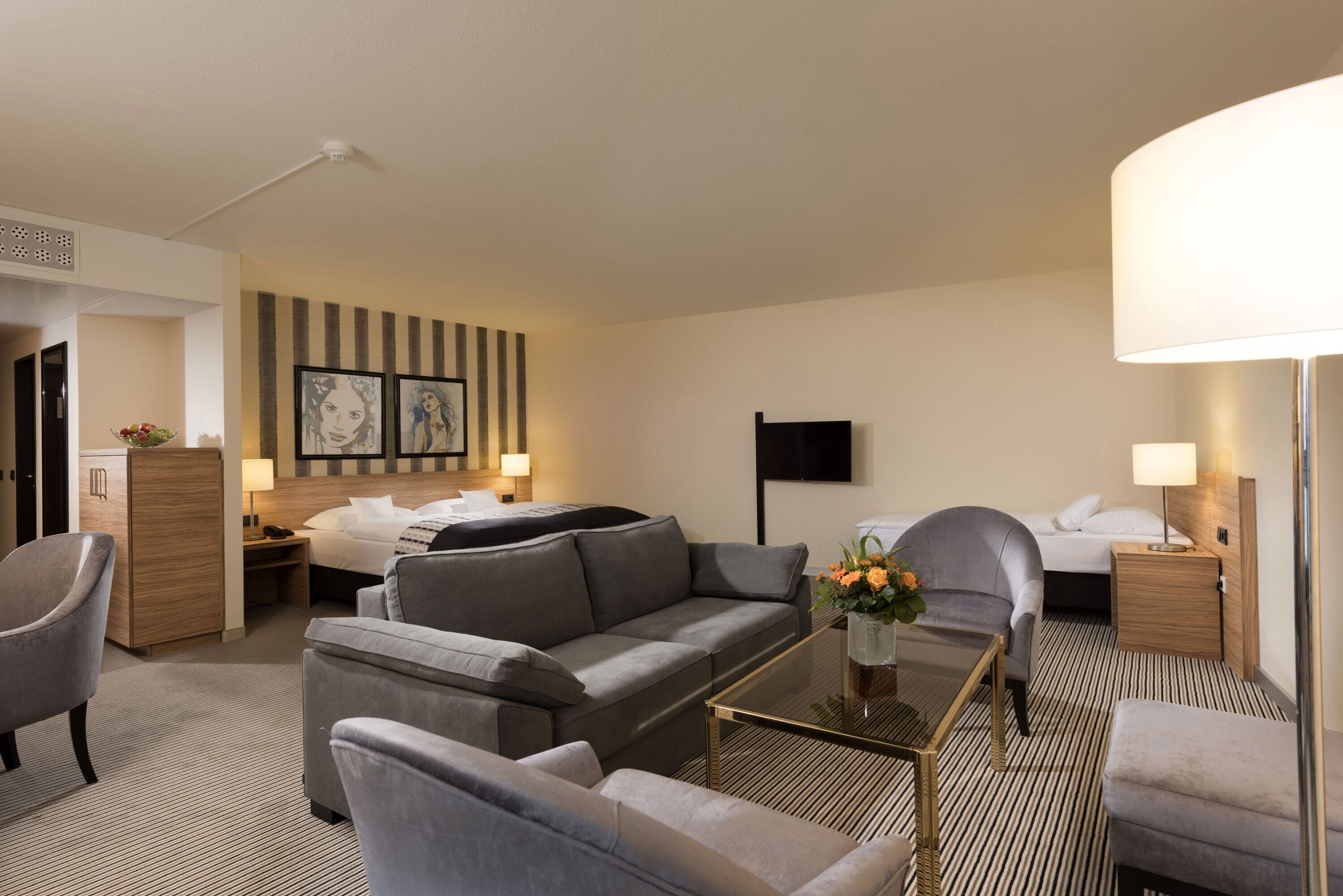 A hotel room with a sofa, armchairs, a glass coffee table, a bed, a TV, and a desk with a lamp.