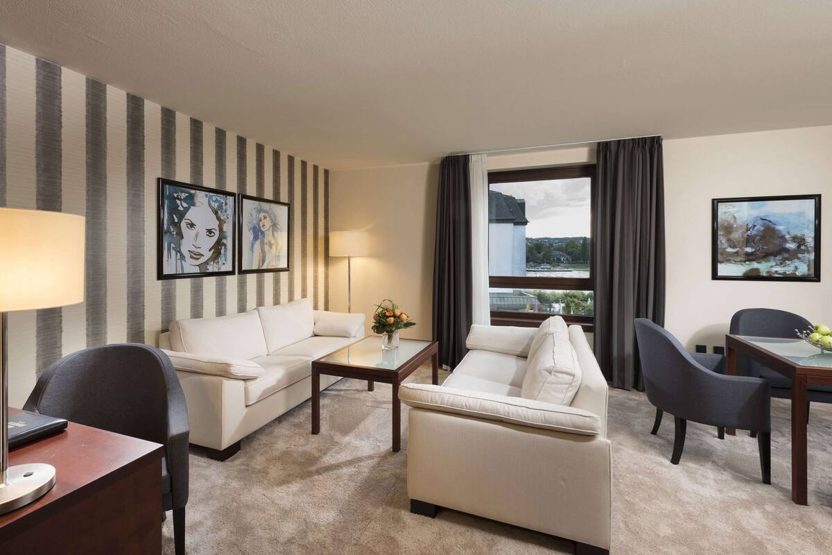 Ein Hotelzimmer mit Streifenmuster-Tapete, zwei weiße Sofas, einem Couchtisch, einem Schreibtisch mit Lampe und Blick ins Freie durch ein Fenster.