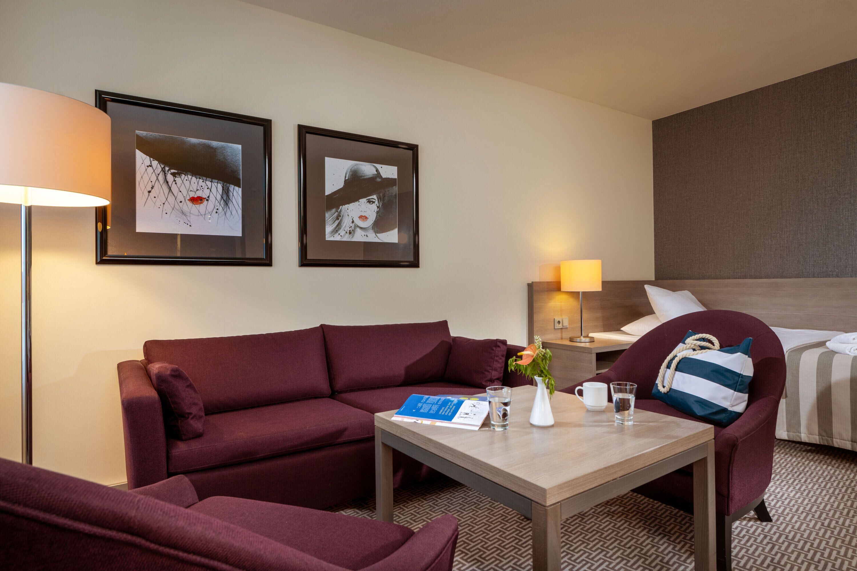 A hotel room with a sofa, a small table with a book, a vase with flowers, a bedside table with a lamp, and a bed with a striped pillow.