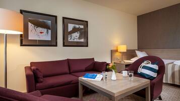 A hotel room with a sofa, a small table with a book, a vase with flowers, a bedside table with a lamp, and a bed with a striped pillow.