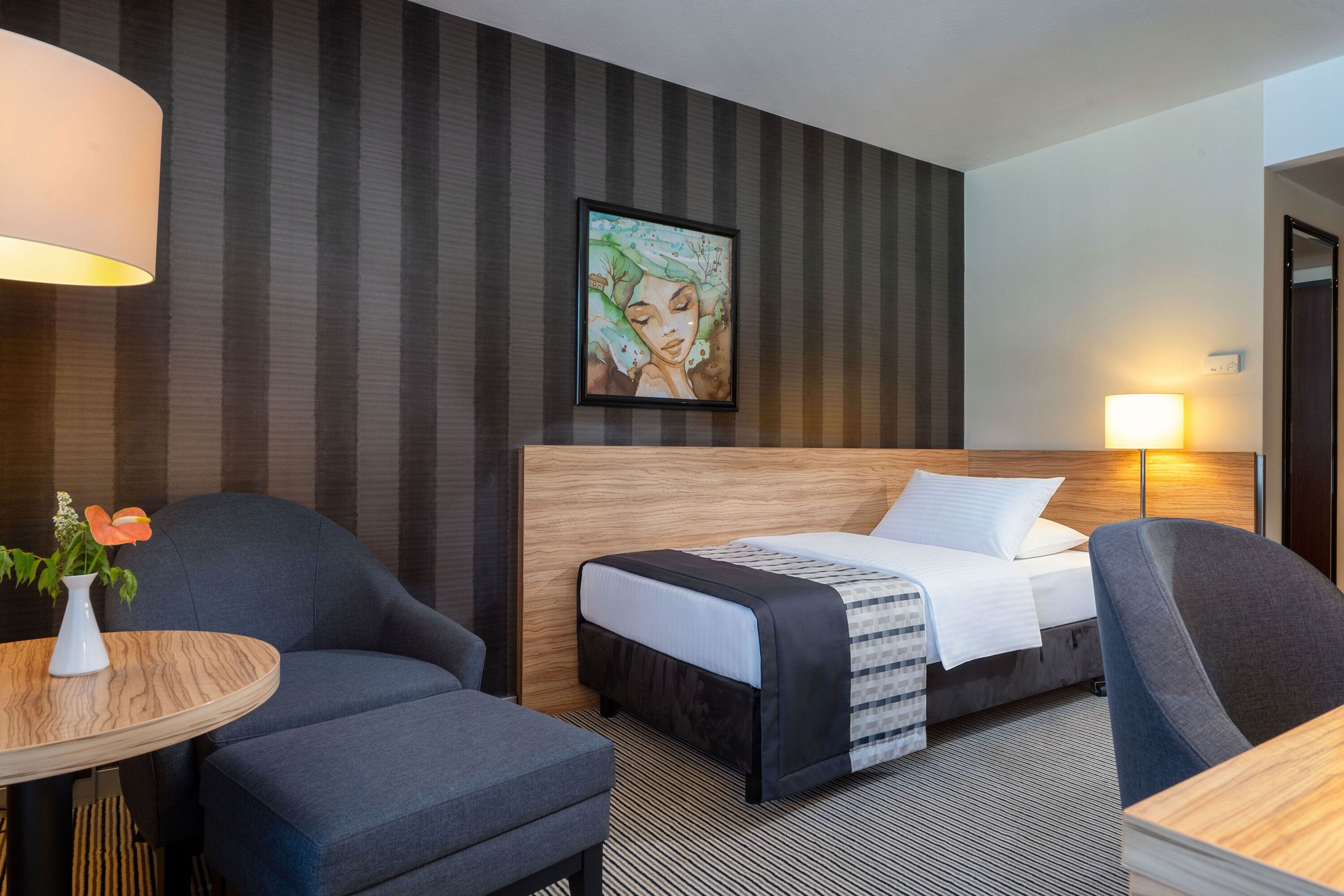 A hotel room with a bed, two armchairs, a small table with a vase, and a painting on the wall.