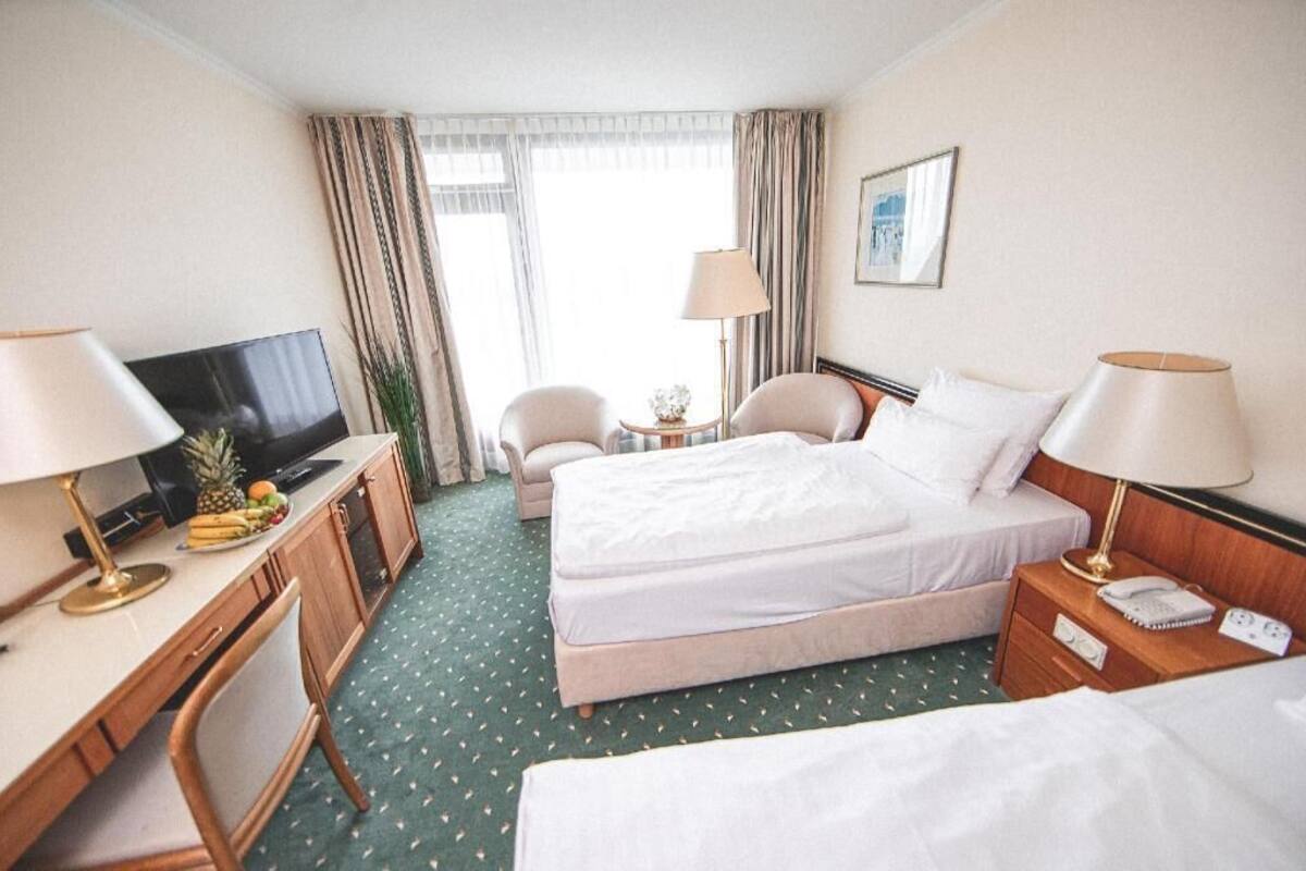 A hotel room with a bed, desk, TV, chairs, and a small table with a lamp.