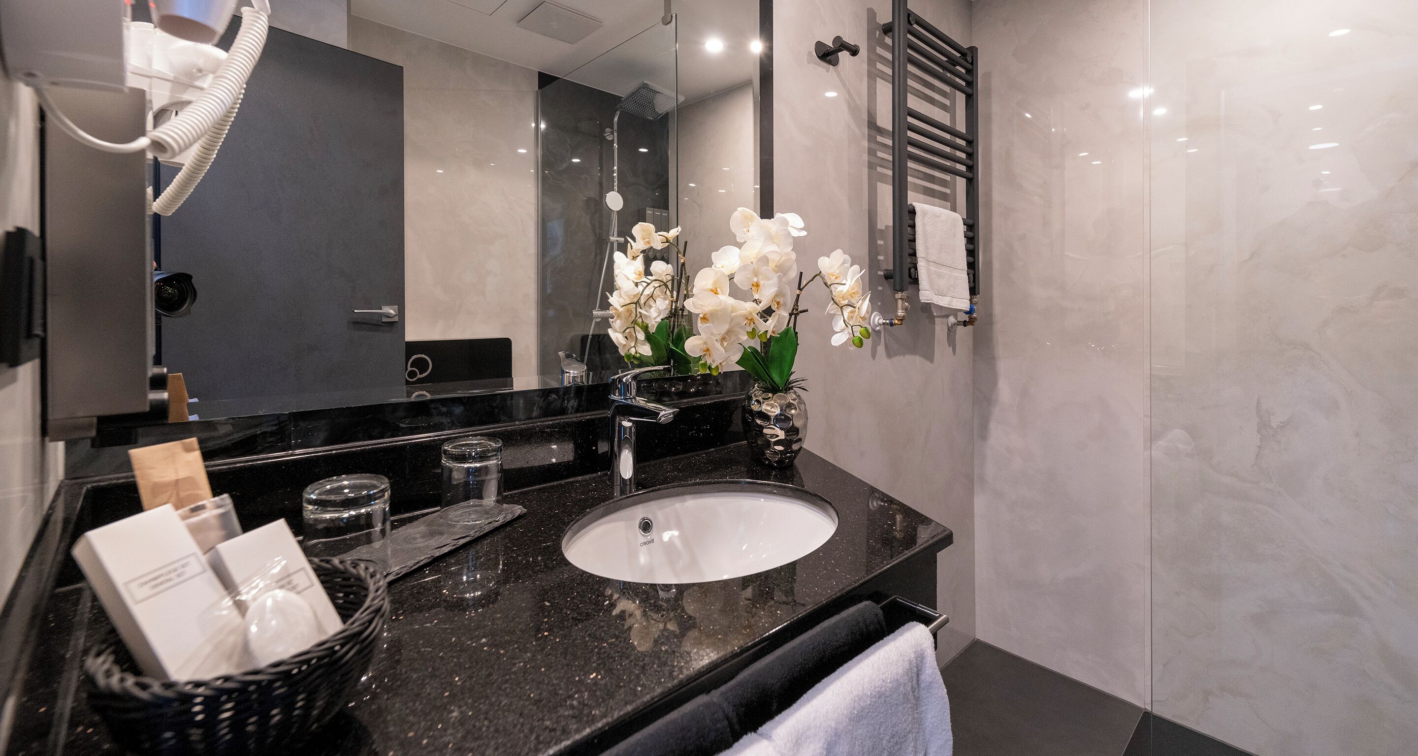 A modern bathroom with a black granite sink, white orchid flowers, a towel rack, and a glass shower enclosure.