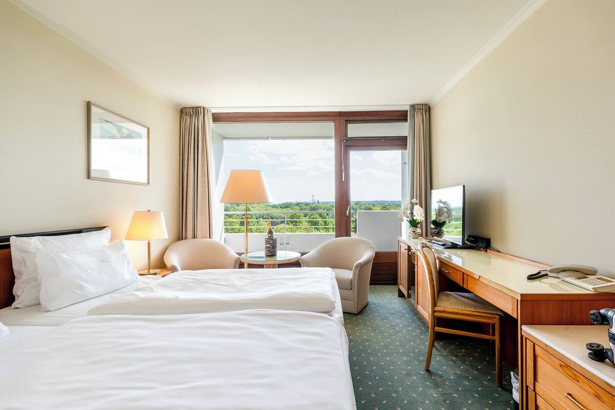 A hotel room with a bed, a desk, a chair, a TV, and a window with a view of greenery.