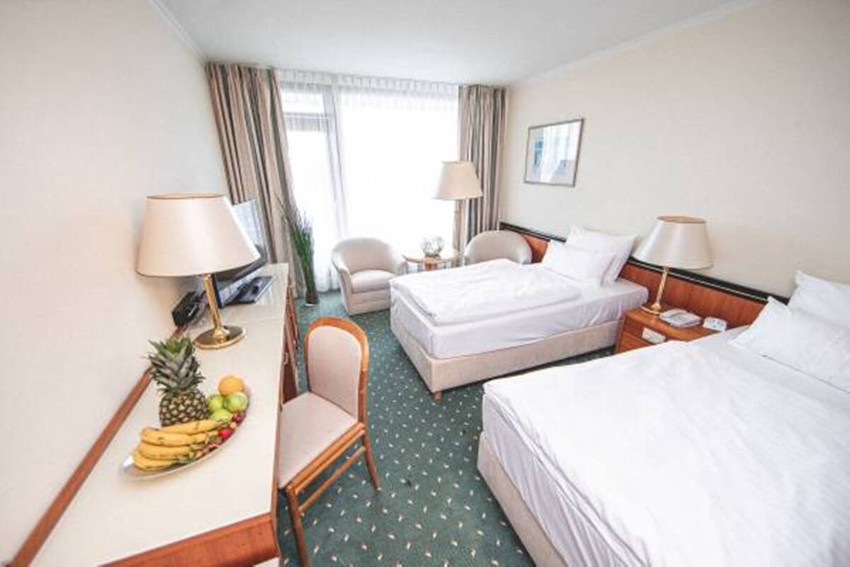 A hotel room with two beds, a desk with a fruit bowl, a chair, and a small table with a lamp.