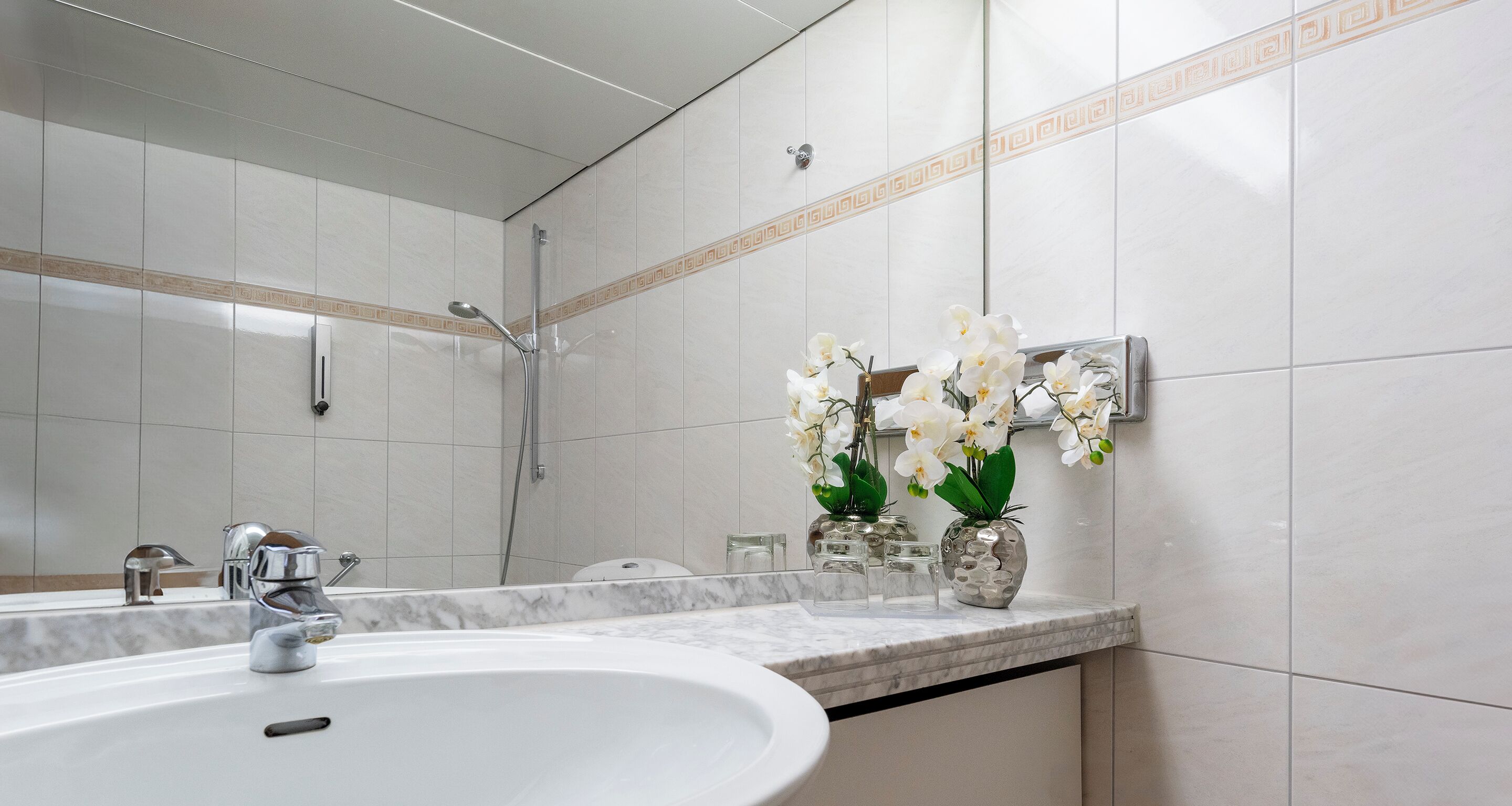 A bathroom with a white sink, a mirror, a shower, and decorative vases with orchids.