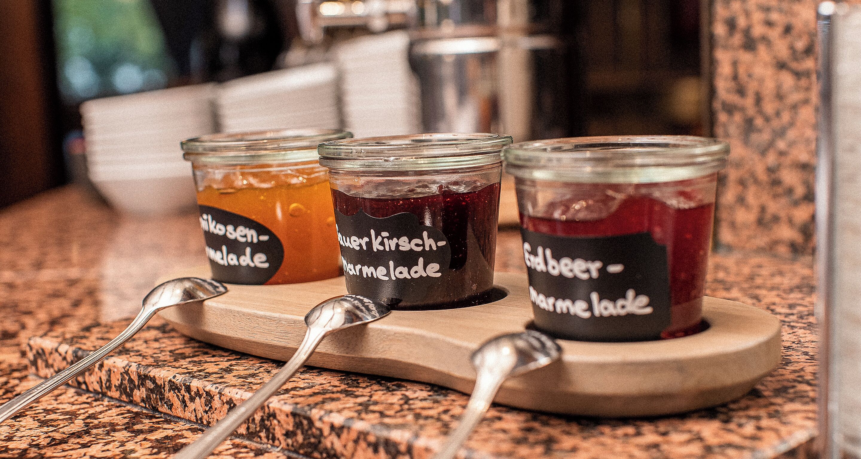 Trois pots en verre, chacun portant une étiquette, sont disposés sur un plateau en bois. Chaque pot contient un type de confiture différent et l’ensemble est placé sur un comptoir en granit.