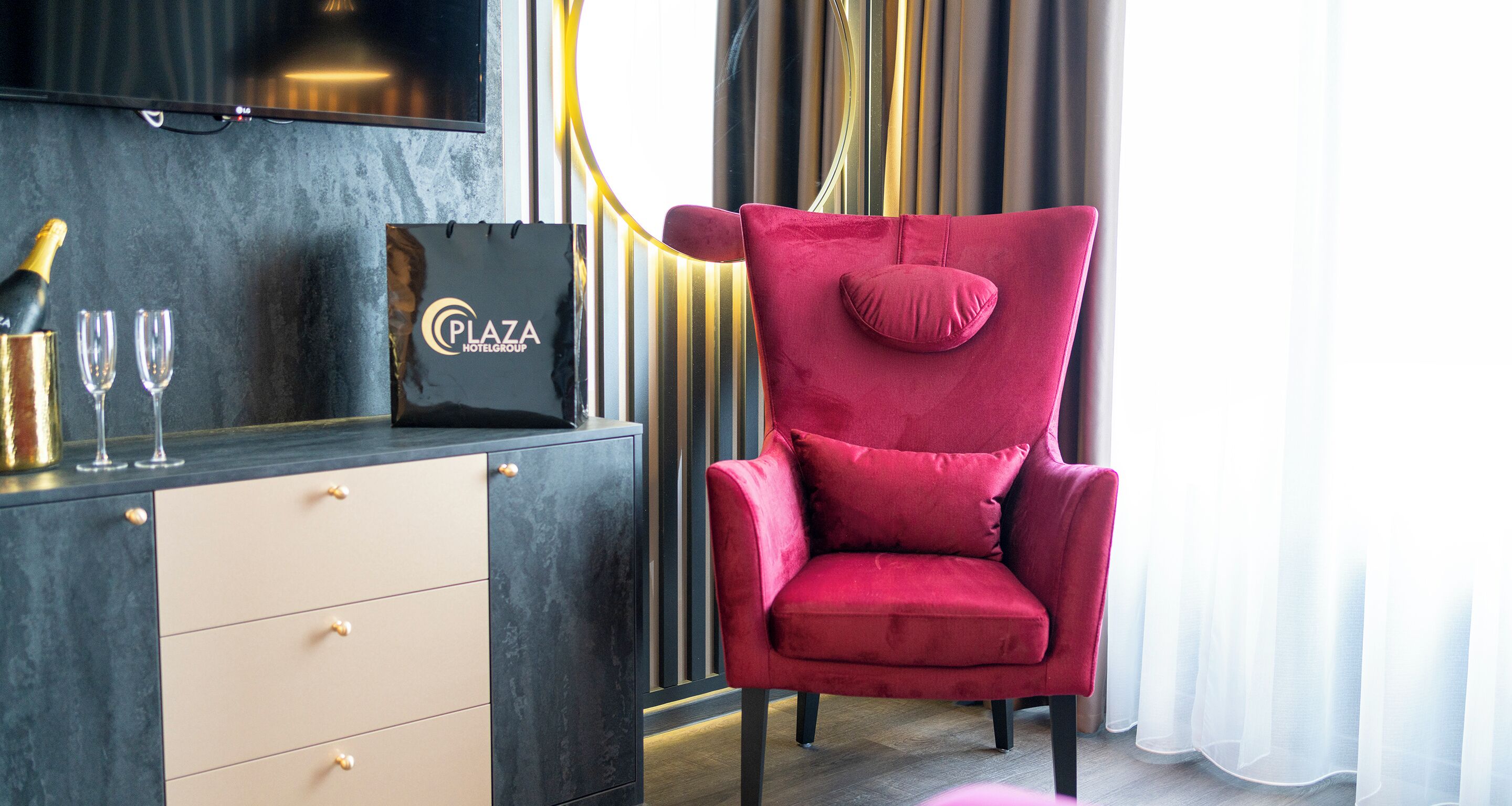 A red velvet chair with a cushion, a minibar with a bottle of champagne and glasses, and a black bag with the logo "Plaza Hotel Group".