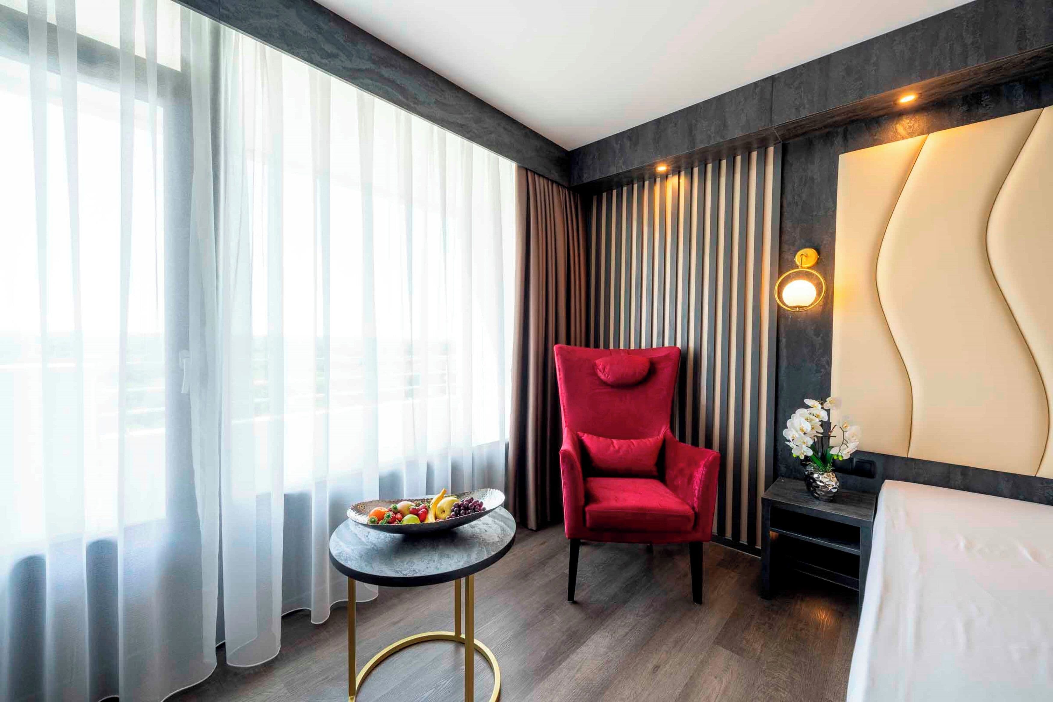 A hotel room with a large window, a red chair, a small round table with a fruit bowl, a bedside table with a vase of flowers, and a bed with a headboard.