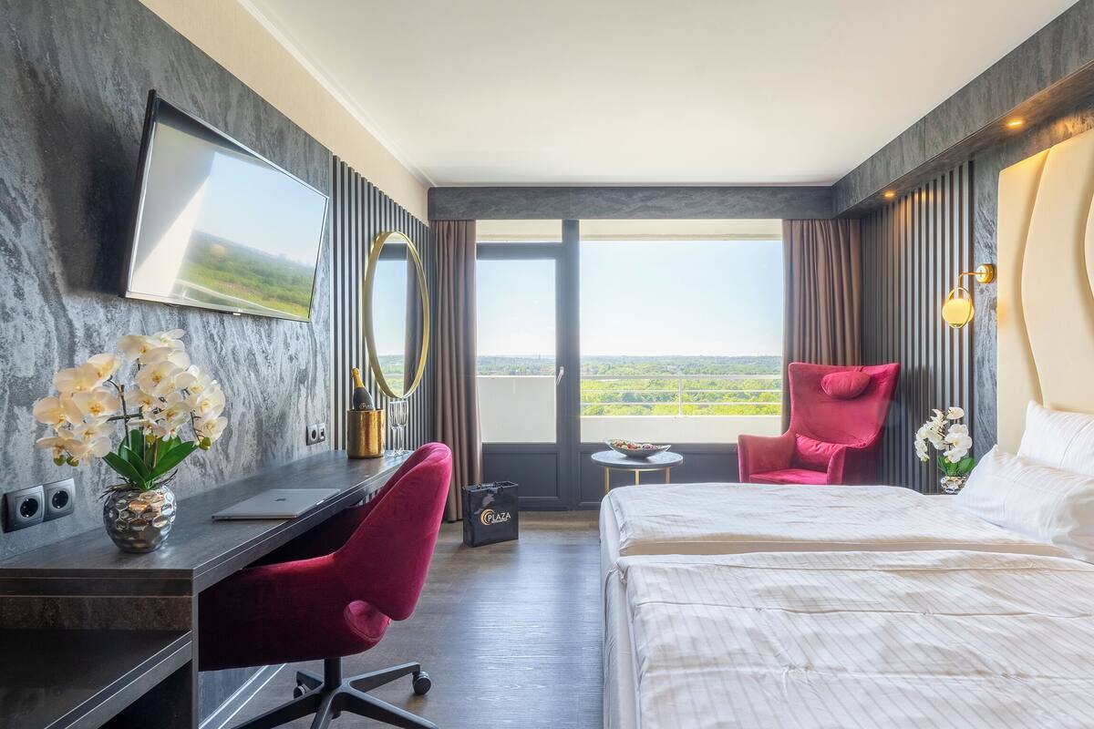 A hotel room with a bed, desk, chair, and a large window with a view.
