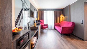 Ein modernes Hotelzimmer mit Holzboden, einer roten Couch, einem kleinen Tisch mit Obstschale, einem Fernseher und einer Minibar.