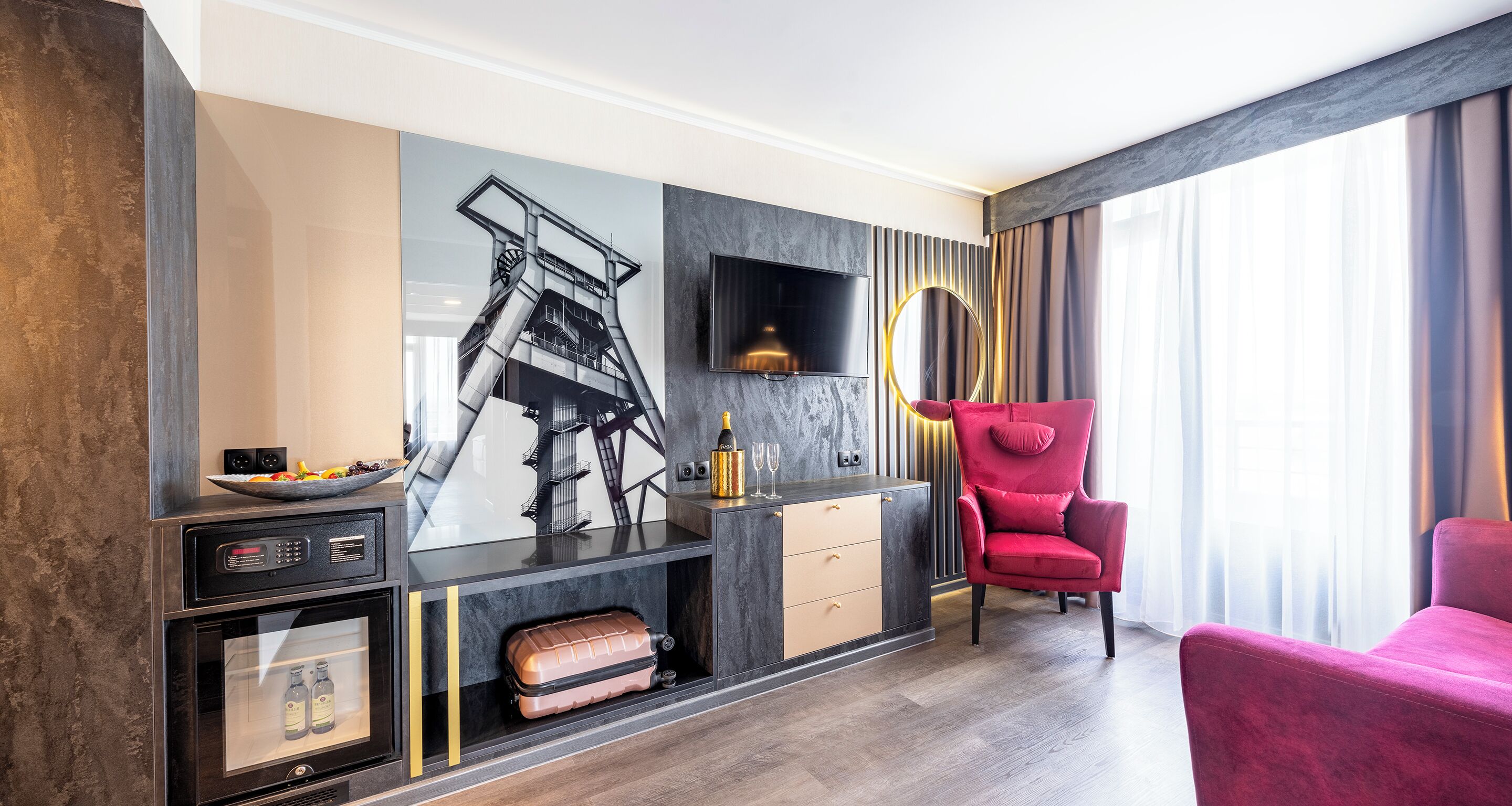 Une chambre d’hôtel moderne dotée d’un grand téléviseur fixé au mur, d’un fauteuil rouge, d’un plancher en bois et d’un mini-bar.