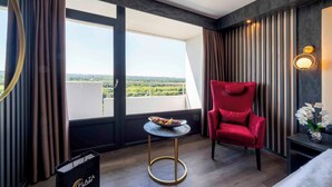 Ein Hotelzimmer mit großem Fenster, einem roten Sessel, einem kleinen runden Tisch und einer Vase mit Blumen.