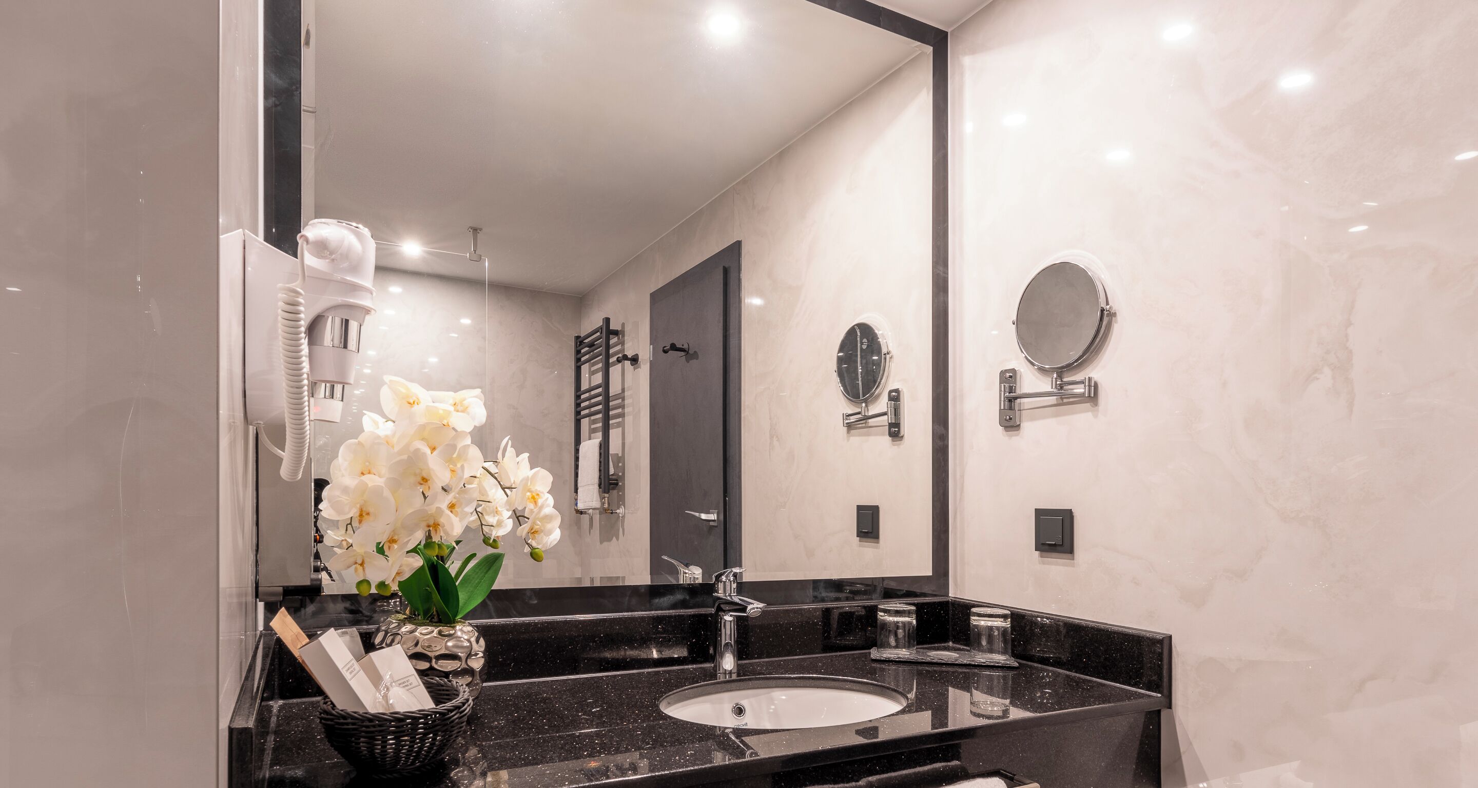 A modern bathroom with a black marble sink, a large mirror, and white orchids.