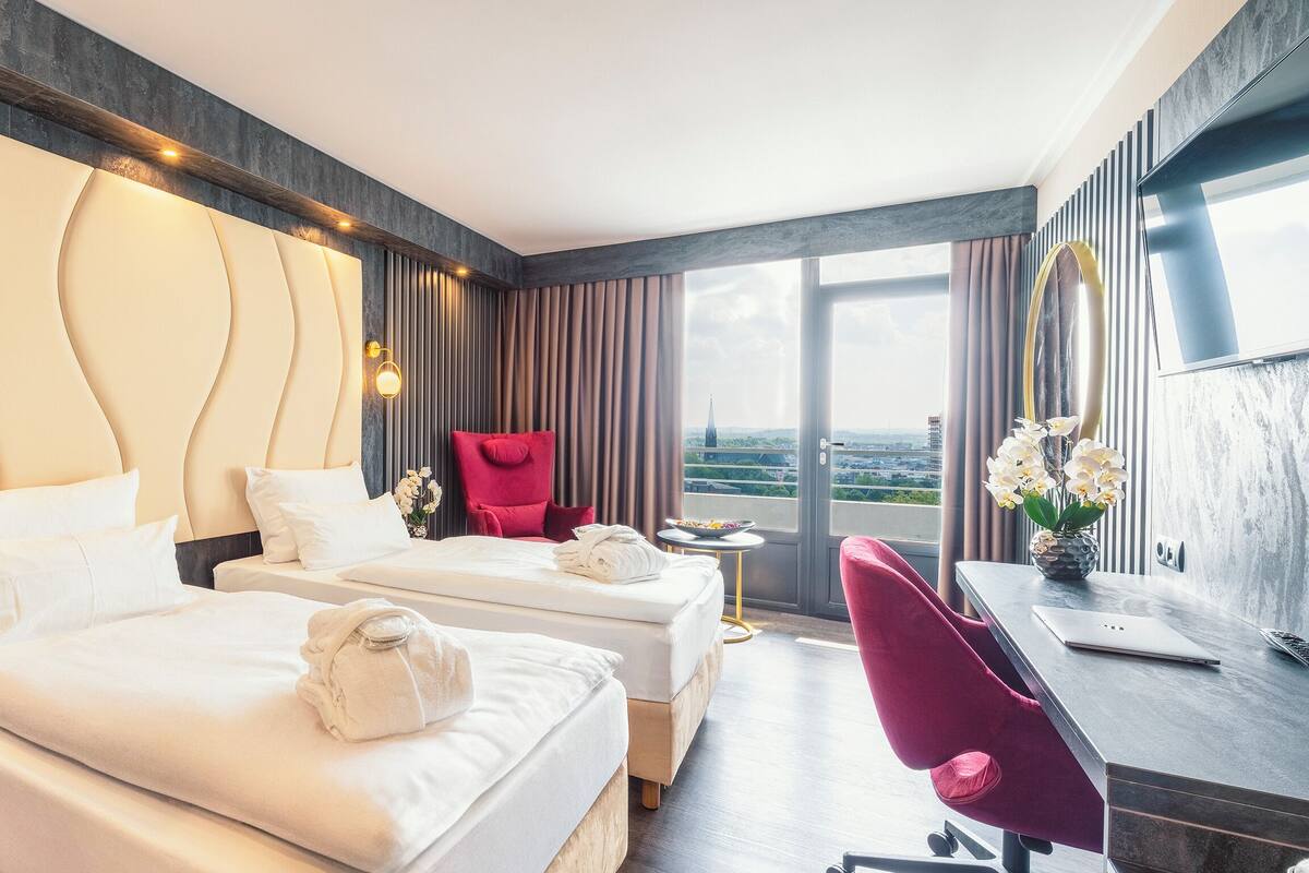 A hotel room with two beds, a desk, a chair, and a large window with a view.