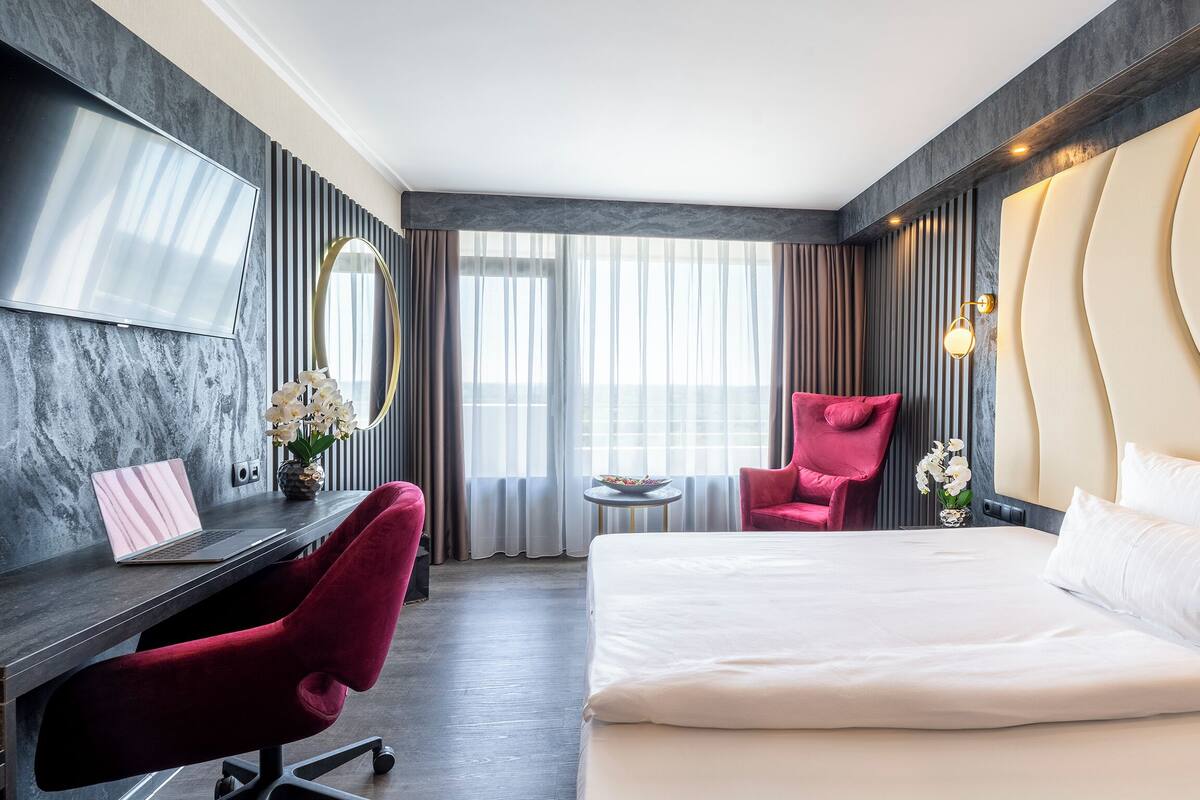 A modern hotel room with a large bed, a desk with a laptop, a red chair, a round table, and a TV mounted on the wall.