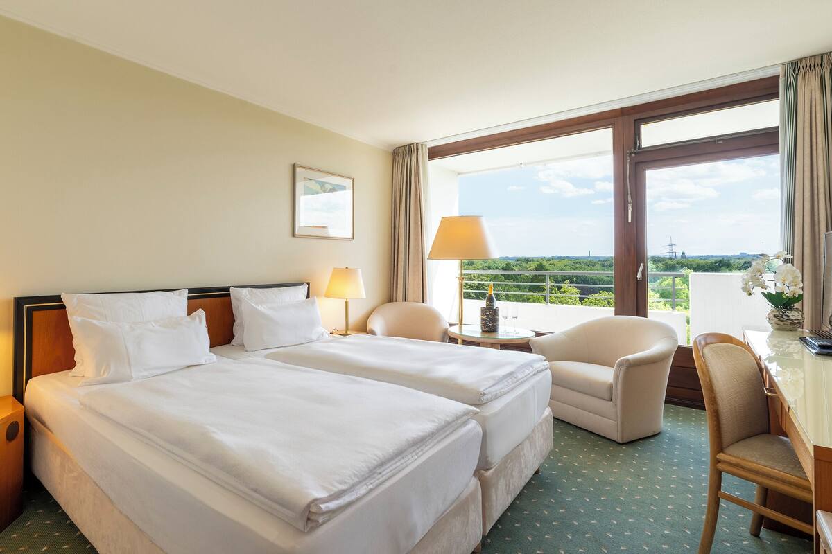 A hotel room with two beds, a desk, a chair, a window with a view, and a lamp on each side of the bed.