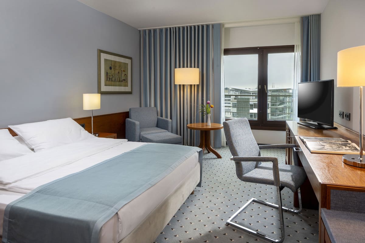 Una habitación de hotel con una cama grande, dos sillones, un escritorio con televisor y un ventanal con vistas a la ciudad.