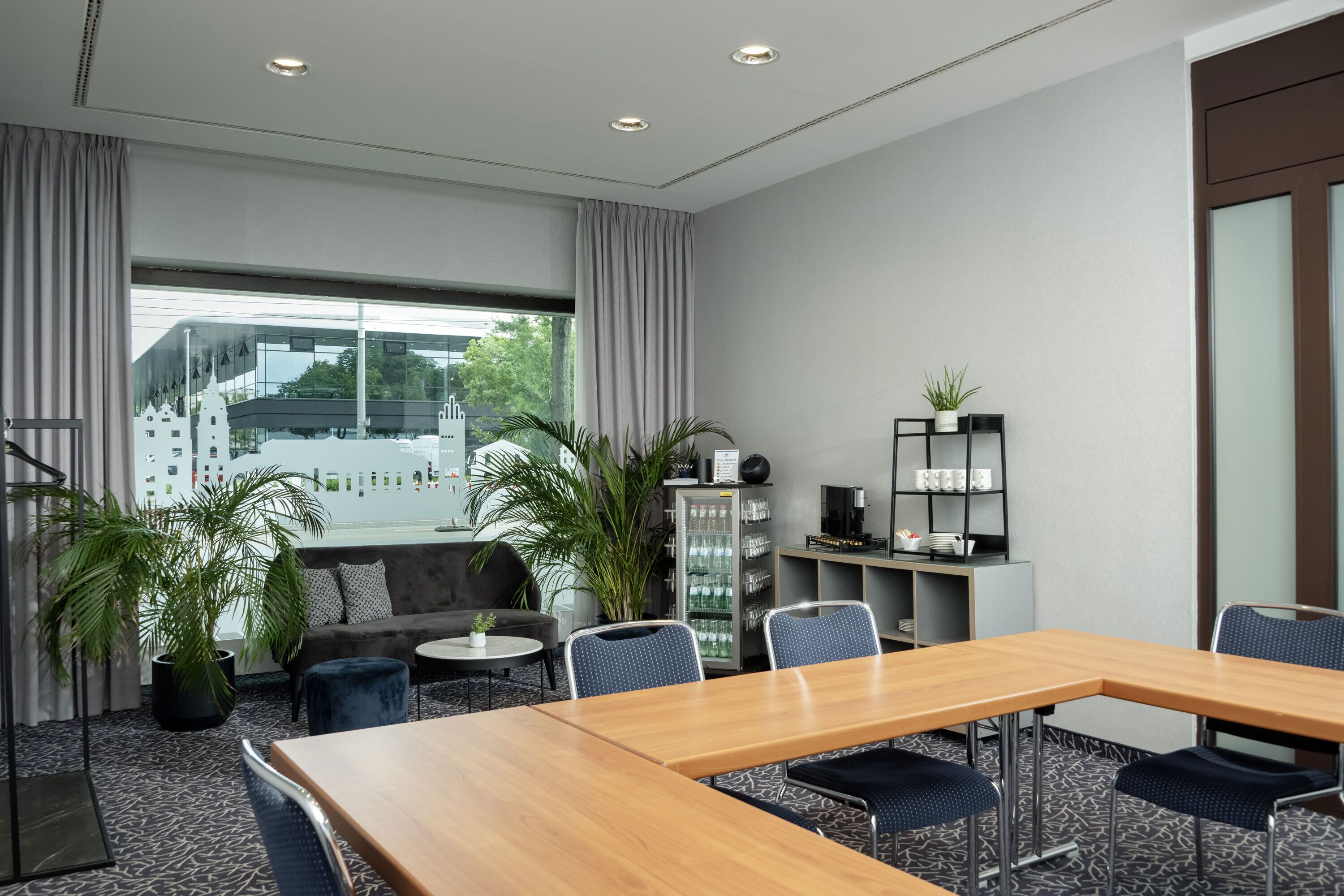 Ein modernes Hotelzimmer mit einem großen Holztisch, bequemen Sitzgelegenheiten, einer Couch, einem kleinen Couchtisch, Zimmerpflanzen, einer Minibar und Blick auf eine Stadtlandschaft durch große Fenster.