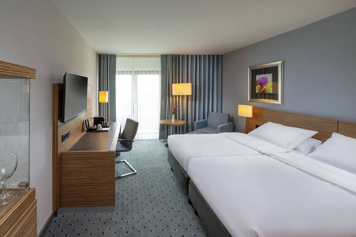 Habitación de hotel con una cama grande, un escritorio con televisor, una silla, una mesita con lámpara y un cuadro en la pared.