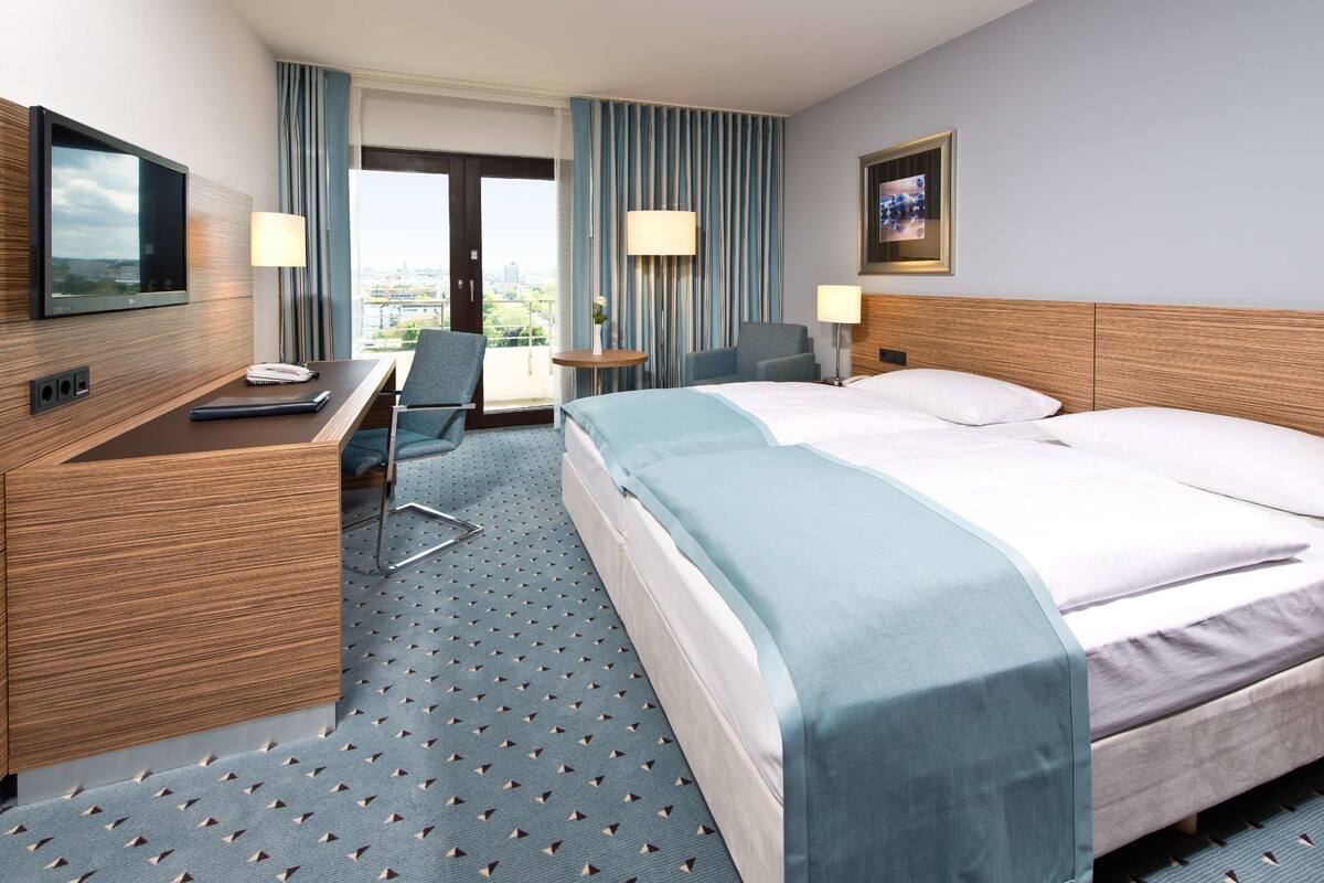 Habitación de hotel con cama, escritorio, silla, televisión y ventana con cortinas.
