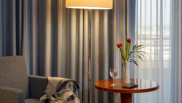 Una habitación de hotel con sofá, una mesa redonda de madera con un florero y un vaso de agua, y una lámpara.